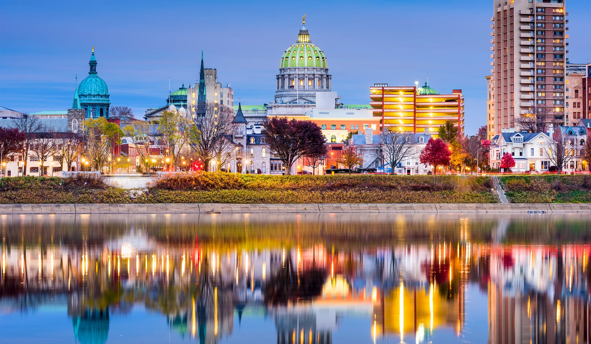 City skyline at dusk, featuring a historic dome, modern buildings, and colorful autumn trees reflected in the water